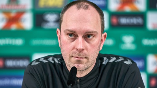 Werder-Trainer Ole Werner sitzt auf dem Podium vor einer grünen Werbewand bei einer Pressekonferenz.