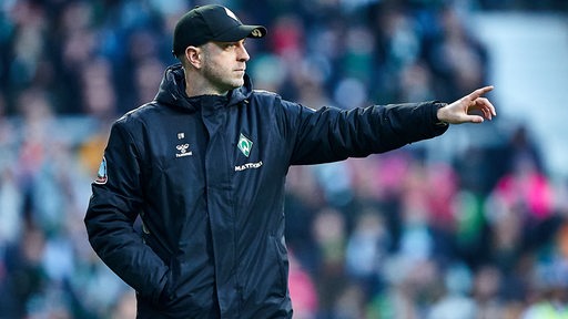 Werder-Trainer Ole Werner zeigt seiner Mannschaft am Spielfeldrand mit ausgestrecktem Finger etwas an.