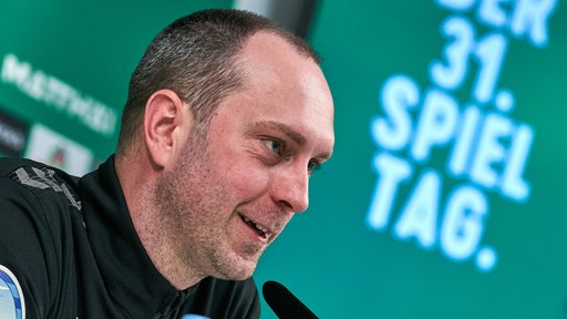 Werder-Trainer Ole Werner strahlt auf dem Podium einer Pressekonferenz.