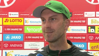 Werder-Trainer Ole Werner steht nach dem Sieg in Mainz lächelnd beim Interview vor einer Werbewand.