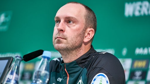 Werder-Trainer Ole Werner sitzt nachdenklich bei einer Pressekonferenz.