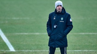 Werder-Trainer Ole Werner steht mit den Händen in der Tasche und einer grauen Wollmütze auf dem Kopf alleine auf dem Trainingsplatz.