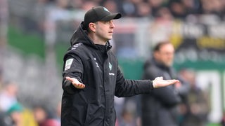 Werder-Trainer Ole Werner steht an der Seitenlinie und macht mit ausgebreiteten Armen eine fragende Geste.