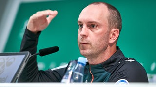 Werder-Trainer Ole Werner deutet auf einer Pressekonferenz etwas mit seiner erhobenen Hand an.