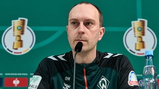 Werder-Trainer Ole Werner sitzt bei einer Pressekonferenz. Im Hintergrund ist das Logo des DFB-Pokals zu sehen.