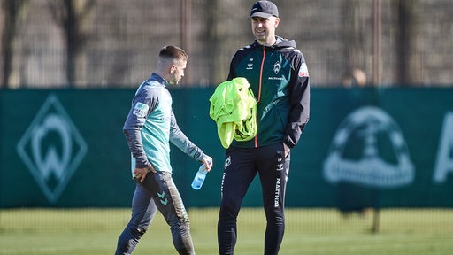 Werder-Trainer Ole Werner steht lächelnd am Rande des Trainings mit einem gelben Leibchen in der Hand, während Romano Schmid an ihm vorbei geht.