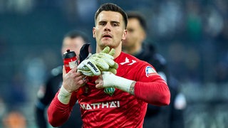 Werder-Torwart Michael Zetterer trägt ein rotes Trikot und schaut nachdenklich nach einem Spiel, während er sich die Handschuhe abstreift.