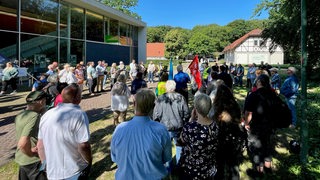 Rund 70 Menschen stehen in einem Park und protestieren.