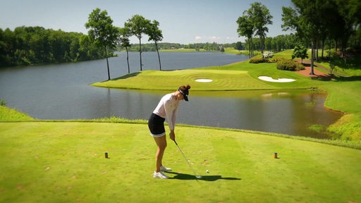 Eine Golferin beim Abschlag auf dem Grün, im Hintergrund ein Gewässer.