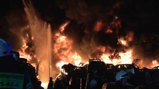 Feuerwehrmänner löschen einen Haufen Autoschrott, welcher in Flammen steht
