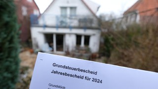 Ein Grundsteuerbescheid für 2024 wird vor einem Privatgrundstück gehalten. 