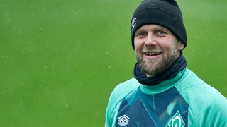 Werder-Stürmer Niclas Füllkrug mit schwarzer Wollmütze auf dem Kopf strahlt am Rande des Trainings.