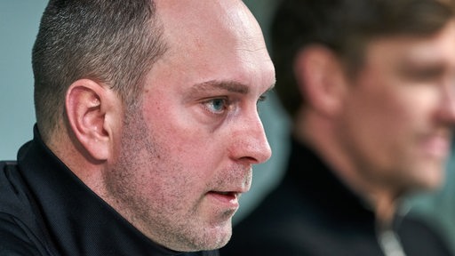 Werder-Trainer Ole Werner von der Seite in Nahaufnahme auf dem Podium einer Pressekonferenz, neben ihm in der Unschärfe Clemens Fritz.