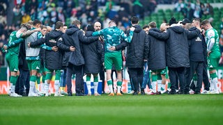 Werder-Spieler und das Trainer-Team stehen nach der Niederlage gegen Freiburg Arm im Arm auf dem Rasen im Kreis.