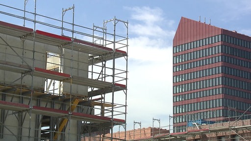 Ein Gerüst an einem Neubau. Rechts daneben ein anderes, hohes Gebäude.