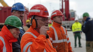 Hafenarbeiter und Lademeister mit Helmen stehen im Hafen, im Hintergrund Container und ein Ladekran.