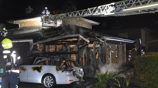 Ein Feuerwehrmann blickt auf ein abgebranntes Haus, vor dem ein stark beschädigtes Auto steht.