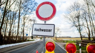 Eine Absperrbake und ein Schild mit der Aufschrift "Hochwasser" steht vor einer gesperrten Straße.