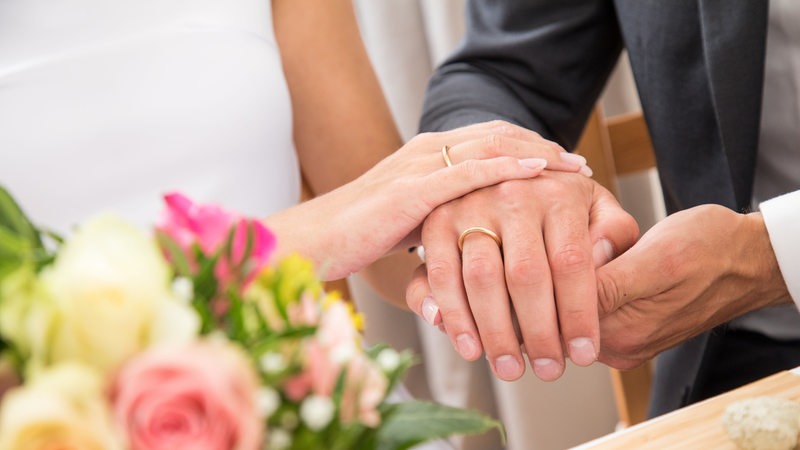 Nahaufnahme von Händen einer Frau und eines Mannes mit Ringen an den Ringfingern. Im Vordergrund ist ein Blumenstrauß zu sehen.