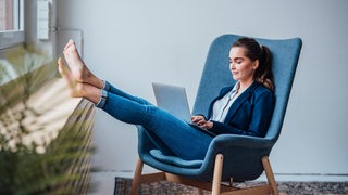 Eine Frau sitzt in einem Sessel am Fenster. Sie hat die Füße hochgelegt und arbeitet an einem Laptop.