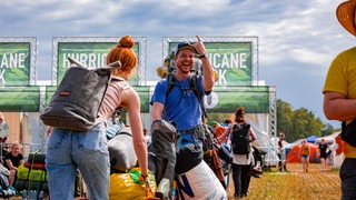 Festivalbesucher lachen vor dem Eingang des Hurricane-Festivals in die Kamera und zeigen eine Rock-Geste.