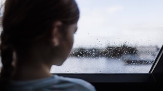 Ein Kind steht vor einem Fenster, auf dem Regentropfen sind (Symbolbild)