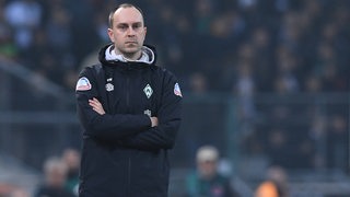 Werder-Trainer Ole Werner steht mit verschränkten Armen und leerem Blick am Spielfeldrand.