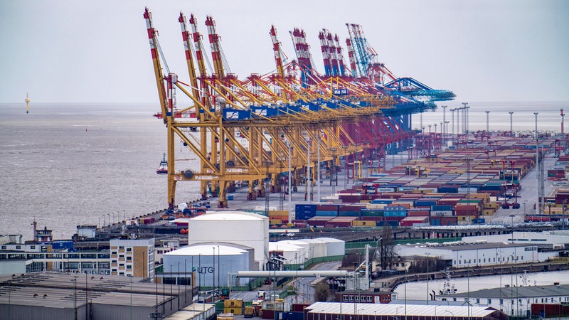 Blick auf mehrere Gebäude, Container und Krananlagen am Wasser.