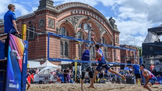 Blick auf ein Beachvolleyball-Feld vor dem Bremer Hauptbahnhof, zwei Spieler springen am Netz hoch.