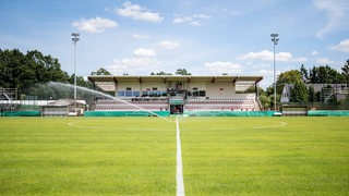 Die Tribüne des Stadions am Vinnenweg ist zu sehen. Ein Rasensprenger bewässert den Fußballplatz.