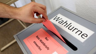 Eine Hand wirft einen Umschlag in eine Wahlurne.