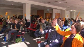 Menschen in einem Saal heben orangene Stimmkarten