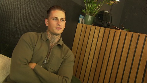 Tattoo-Influencer Fabien Tietjen sitzt auf einem Sofa vor einer schwarzen Wand.