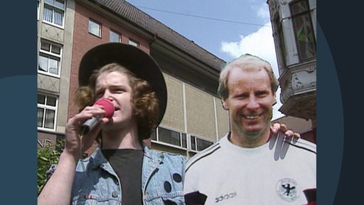 Ein Passant hält ein Mikro in der Hand. Er hält dabei die Pappfigur von Berti Vogts im Arm.