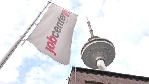 Die Jobcenterflagge neben dem Waller Fernsehturm. Im Hintergrund ein wolkenreicher Himmel.