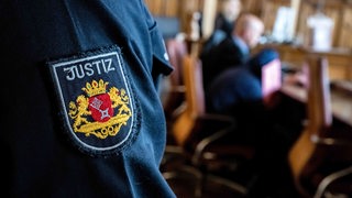 Ein Justizbeamter steht im Gerichtssaal, während der Angeklagte vor Prozessbeginn sein Gesicht hinter einer Mappe verbirgt. 