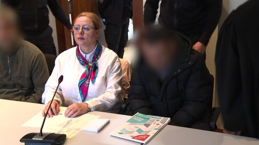 Eine Frau sitzt neben einem Mann an einem Tisch, das Gesicht des Mannes ist zensiert.