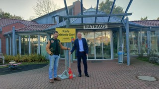 Zwei Männer stehen mit Spaten und Schild vor einem Gebäude.