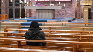 Eine Person in Winterkleidung sitzt in einer Kirchenbank und blickt zum Altar.