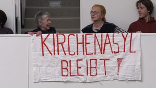 Drei Personen stehen hinter einem Protestbanner mit der Aufschrift Kirchenasyl bleibt!