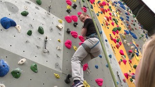 Ein Junge mit einer Behinderung hängt gesichert an einem Seil an einer Kletterwand.