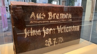 Ein Koffer aus braunem Leder. Seine Kanten sind abgewetzt, man sieht ihm die Jahre an, die er hinter sich hat. Mit weißer Farbe wurde ein Name und eine Adresse drauf gemalt.