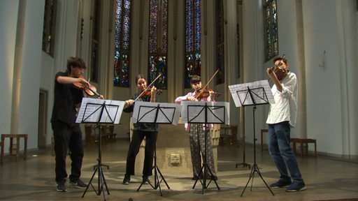 Vier junge Menschen stehen auf der Bühne in einer Kirche und spielen Violine.