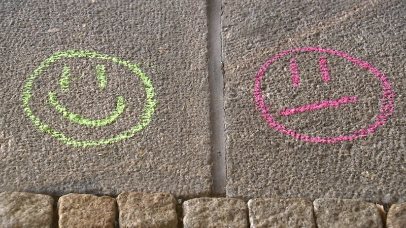 Auf Steinen wurden mit Kreide zwei Smileys gemalt. Einer guckt glücklich und ist grün und einer guckt unglücklich und ist rot.