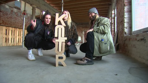 Drei Jugendliche sitzen in einer Halle vor einer Skulptur mit der Aufschrift "Skulptur".