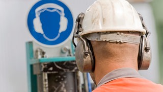 Ein Mann mit Helm und Gehörschutz steht vor einem Gehörschutz-Schild.