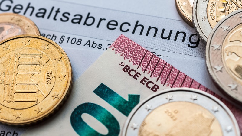 Auf einem Papier mit der Aufschrift "Gehaltsabrechnung" liegen Geldmünzen und eine Euro-Banknote.