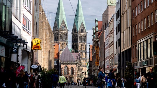 Zahlreiche Menschen sind bei sonnigem Wetter auf der Obernstraße in der Innenstadt unterwegs, während im Hintergrund der historische Bremer Dom zu erkennen ist.