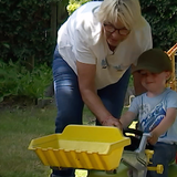 Eine Frau beugt sich zu einem kleinen Jungen auf einem Tret-Trecker herunter und schiebt ihn an.