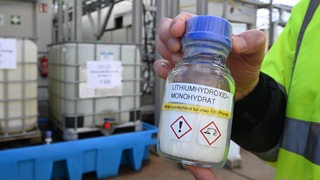 Jemand hält eine Flasche mit weißem, salzartigen Inhalt in die Kamera. Darauf steht: Lithiumhydroxid-Monohydrat.